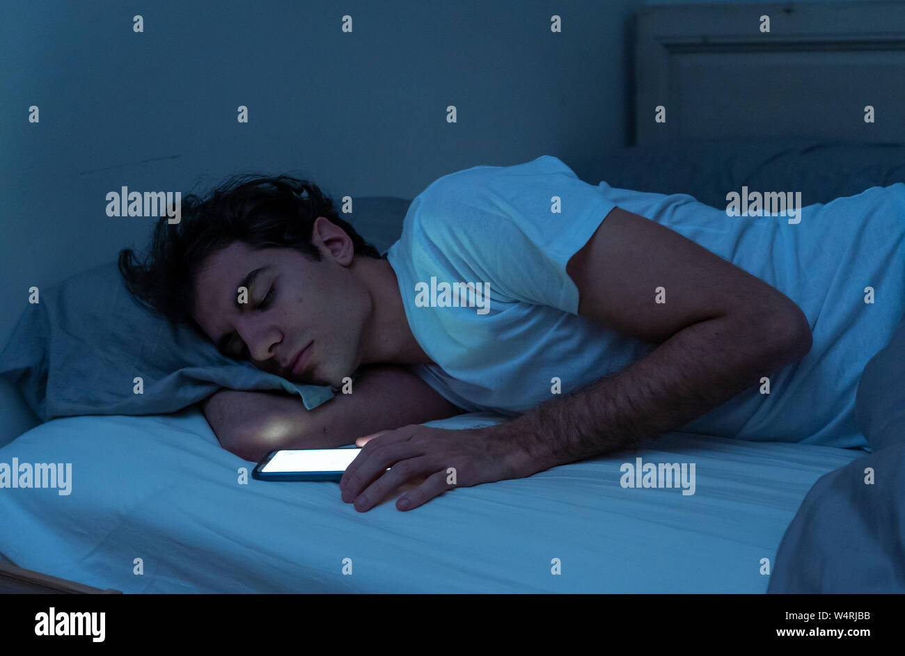 Addicted to social media giovane uomo addormentarsi con smart telefono cellulare durante la notte in letto. lifestyle ritratto dell'uomo dorme nella camera oscura con mob Foto Stock