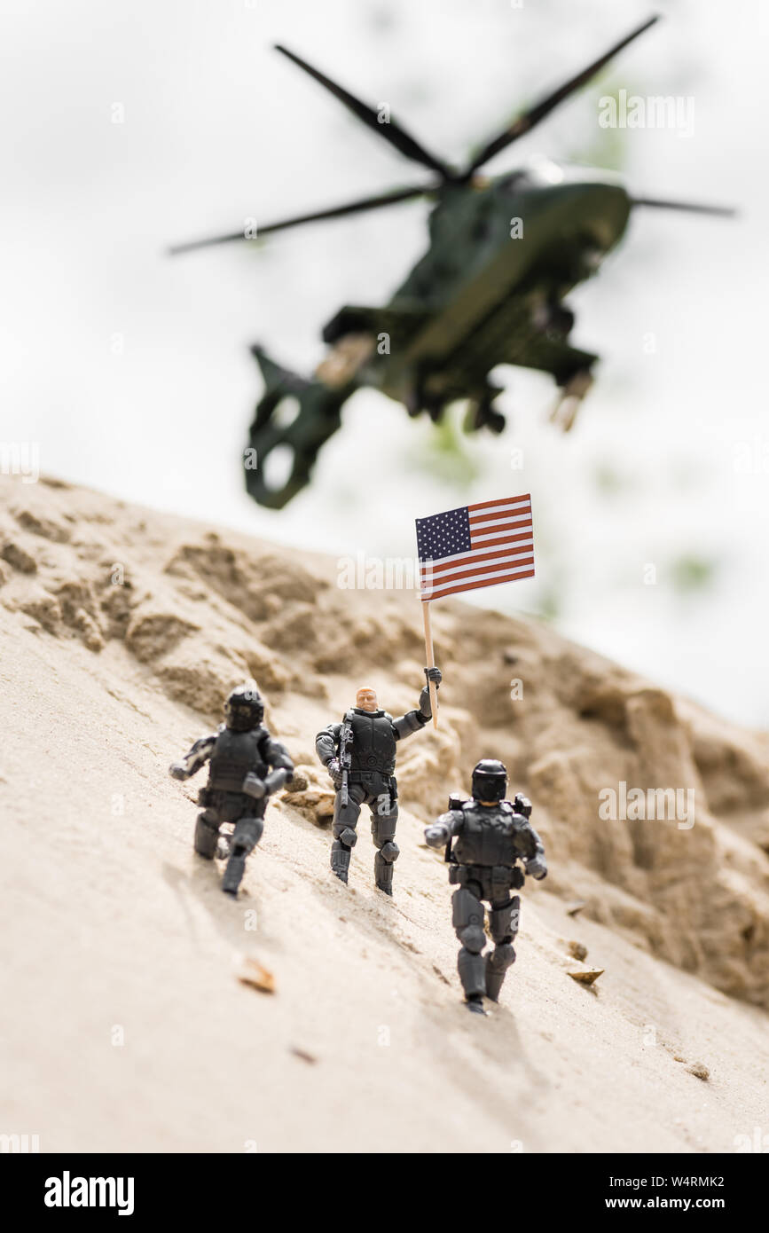 Messa a fuoco selettiva del soldatino con arma holding bandiera americana sulla sabbia con elicottero sullo sfondo Foto Stock