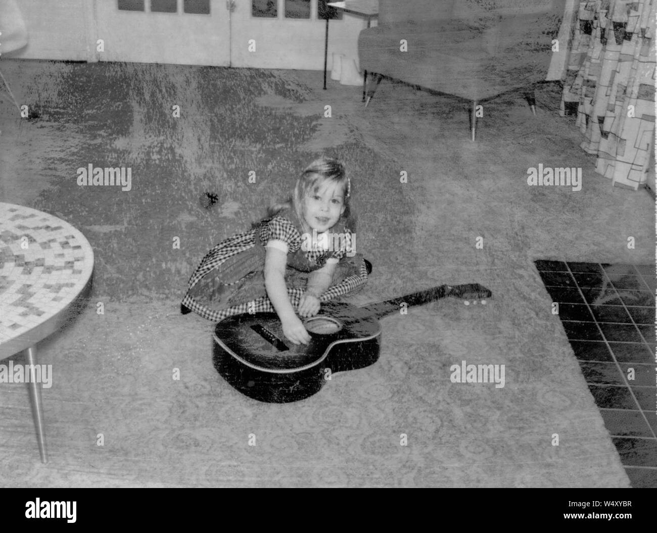 Giovane ragazza abito da indossare, strimpellamento una chitarra che giace sul piano di un soggiorno, con tavolo da caffè, sedia e camino visibile, danni sulla fotografia originale, Detroit, Michigan, 1960. () Foto Stock