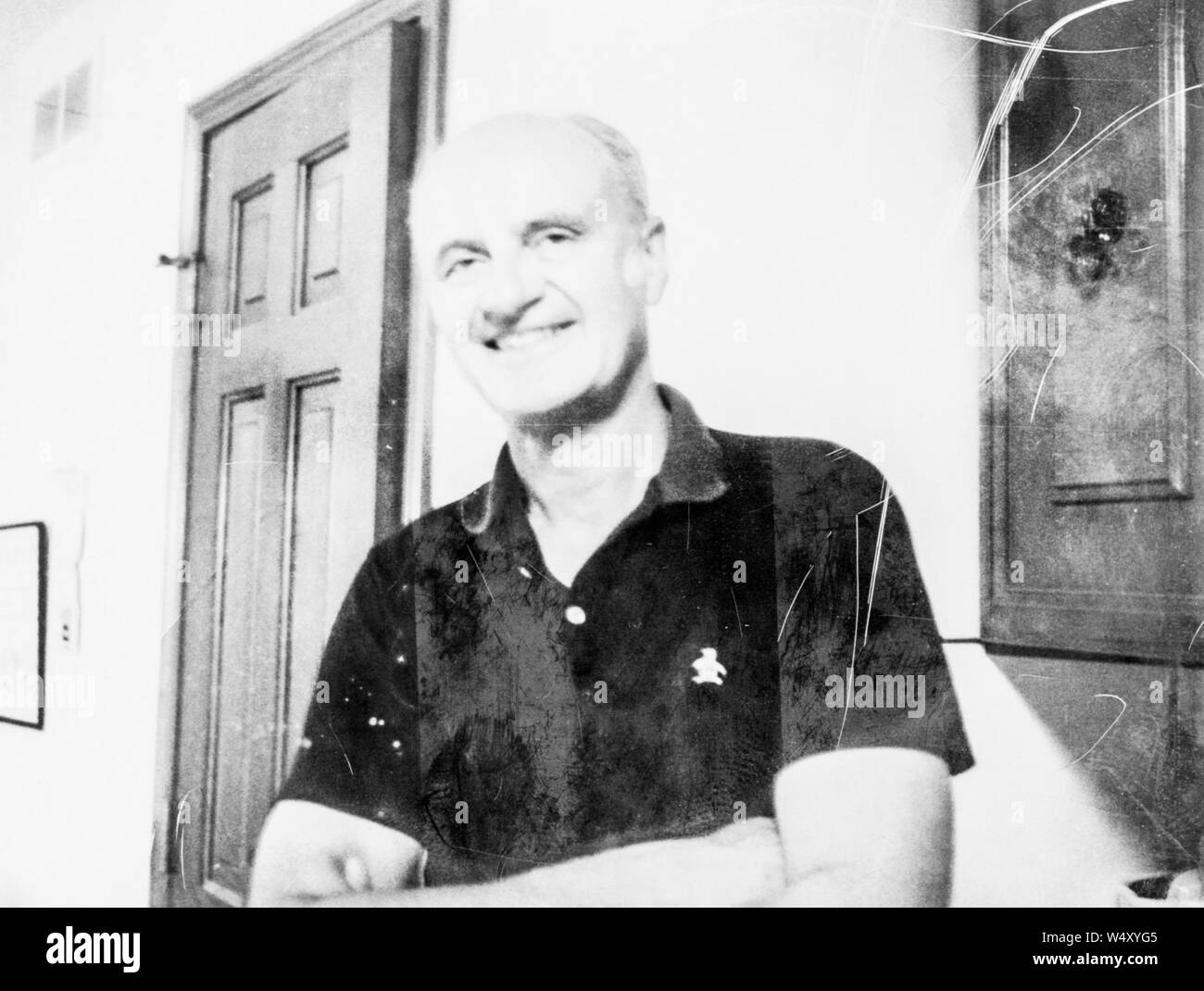 Mezza lunghezza Ritratto di un uomo che indossa una breve sleaved, stile Polo shirt, con le braccia incrociate, sorridere mentre in piedi nel corridoio di una casa a Detroit, Michigan, ingenti danni sulla fotografia originale, 1970. () Foto Stock