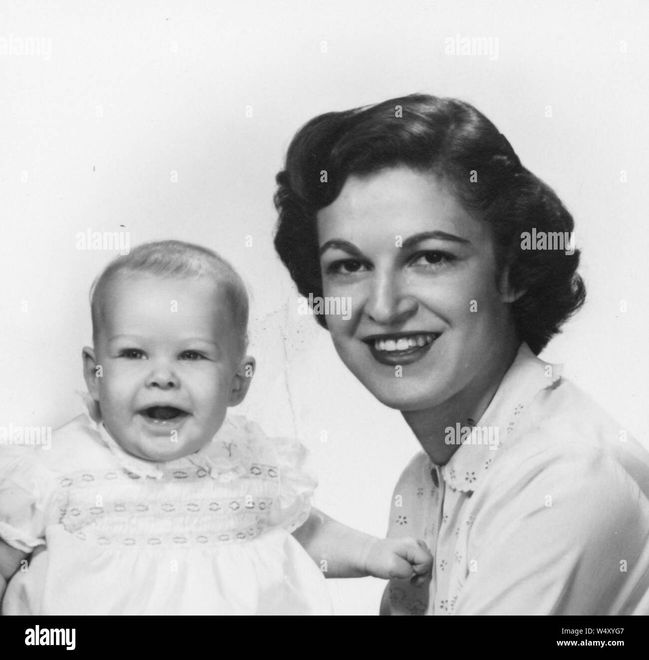 Testa e spalle il ritratto di una giovane madre indossa una camicia bianca, sorridente e tenendo la sua figlia infante, che guarda verso la telecamera con bocca aperta, 1960. () Foto Stock