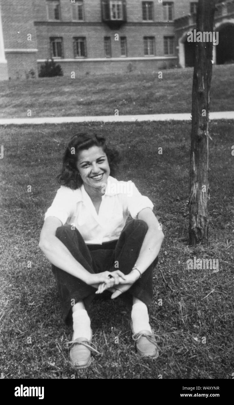 Giovane donna indossa camicia e pantaloni sorridente e seduta sul prato con le braccia che circondano le gambe, 1950. () Foto Stock