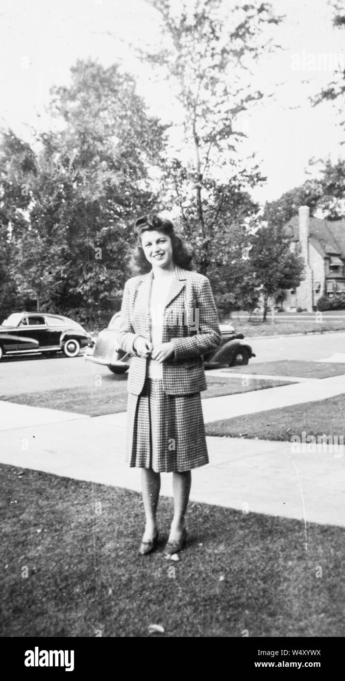 A piena lunghezza Ritratto di giovane donna che indossa a scacchi in giacca e gonna in piedi sul prato anteriore della sua casa, con diverse vetture visibili in background, Detroit, Michigan, 1950. () Foto Stock