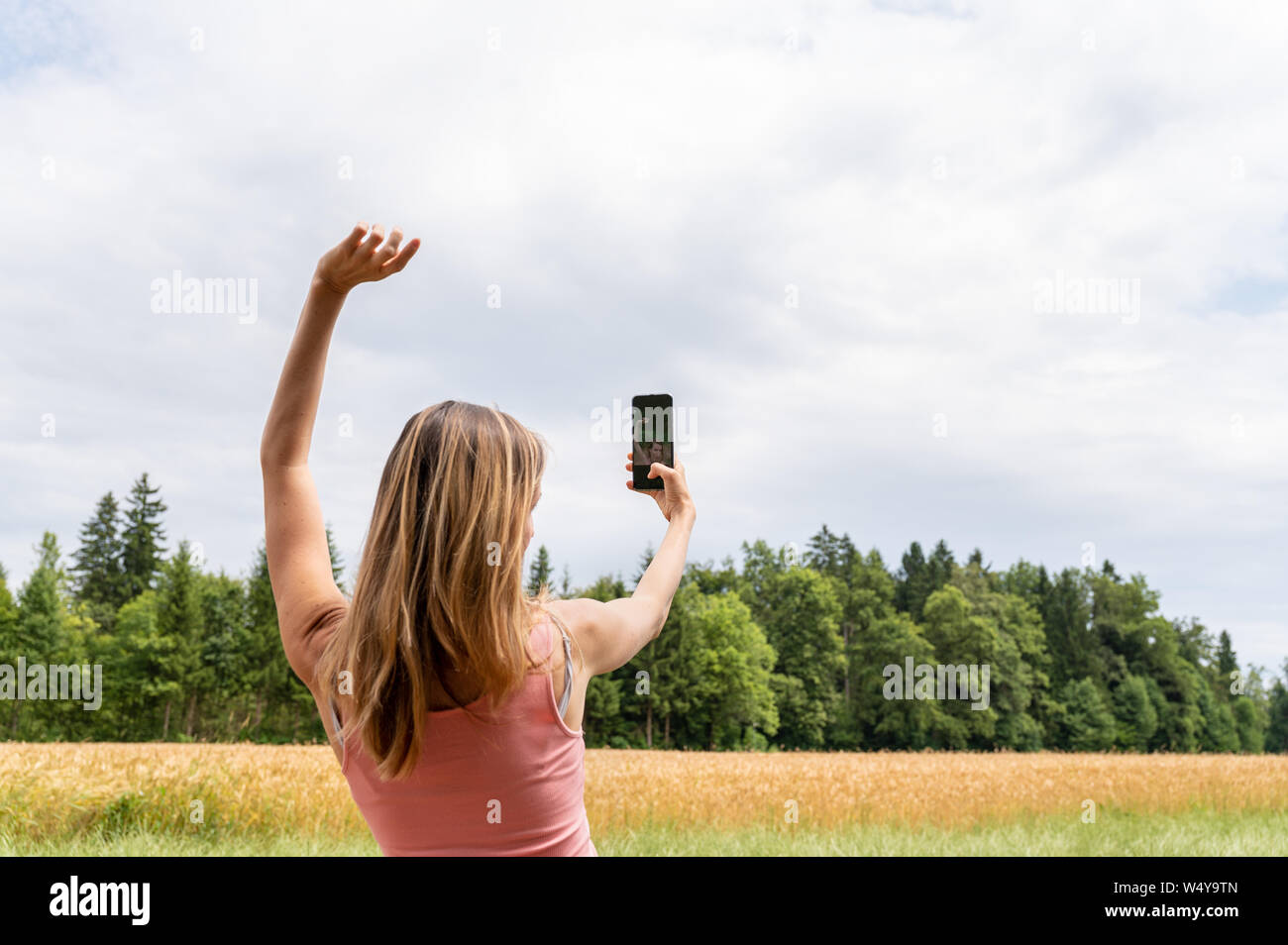 Vista da dietro di una giovane donna che fa selfie con il suo telefono cellulare al di fuori in natura mentre si effettua la posa con il suo braccio nell'aria. Foto Stock