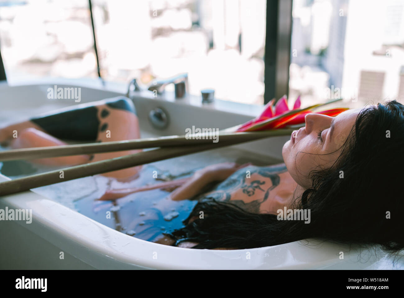 Bruna giovane donna con tatuaggi tenendo bagno con fiore tropicale in bagno luminoso con vista città Foto Stock