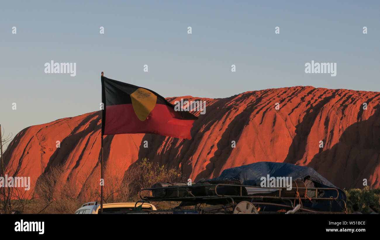 La bandiera aborigena Vola di fronte ofUluru o Ayers Rock in Australia centrale. L'enorme monolito di pietra arenaria è sacro agli indigeni australiani. Foto Stock