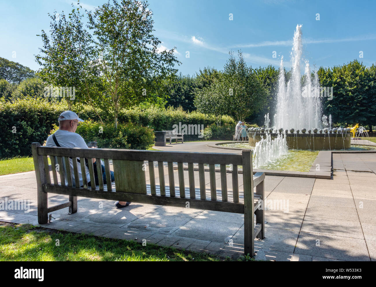 Un uomo si siede su una panchina nel parco presso la fontana del Giubileo in Goswell Park, Windsor, Berkshire, Regno Unito Foto Stock