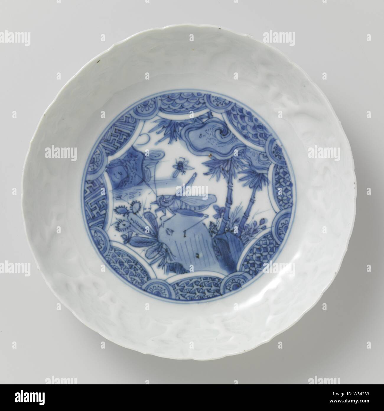 Piatto, piattino - piatto con un grasshopper su una roccia, porcellana piatto con un bordo dentellato, dipinta in underglaze blu. Sul ripiano di una stella cartiglio con una cavalletta su una roccia con piante fiorite e bambù. Il cartiglio igienico il lavoro e la metà dei fiori. Sul bordo interno di una band con vitigni di fiori e melograni in rilievo. La cottura della sabbia sui piedi anello. Porcellana crepata in blu e bianco, insetti: erba-tramoggia, anonimo, Cina, c. 1580 - c. 1620, Ming-dinastia (1368-1644) / Wanli-periodo (1573-1619), porcellana (materiale), smalto, cobalto (minerali), di vetrificazione, h 3,8 cm d 20,1 cm d 12,9 Foto Stock