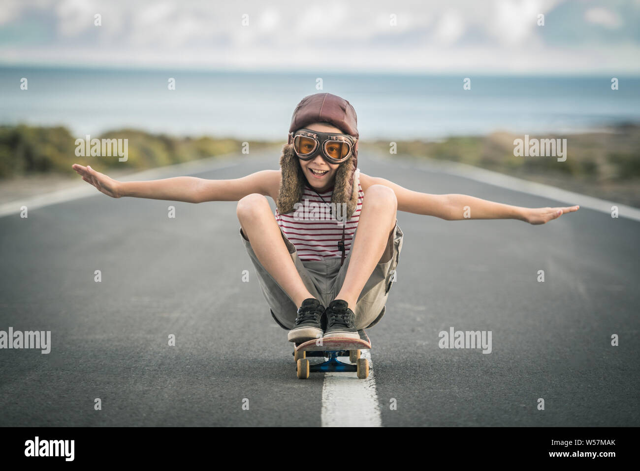 Giovane ragazzo biondo gioca aviatore seduta con le gambe incrociate su  skateboard con braccia tese a volare sorridente bambino imita aereo sulla  pista di aeroporto Foto stock - Alamy