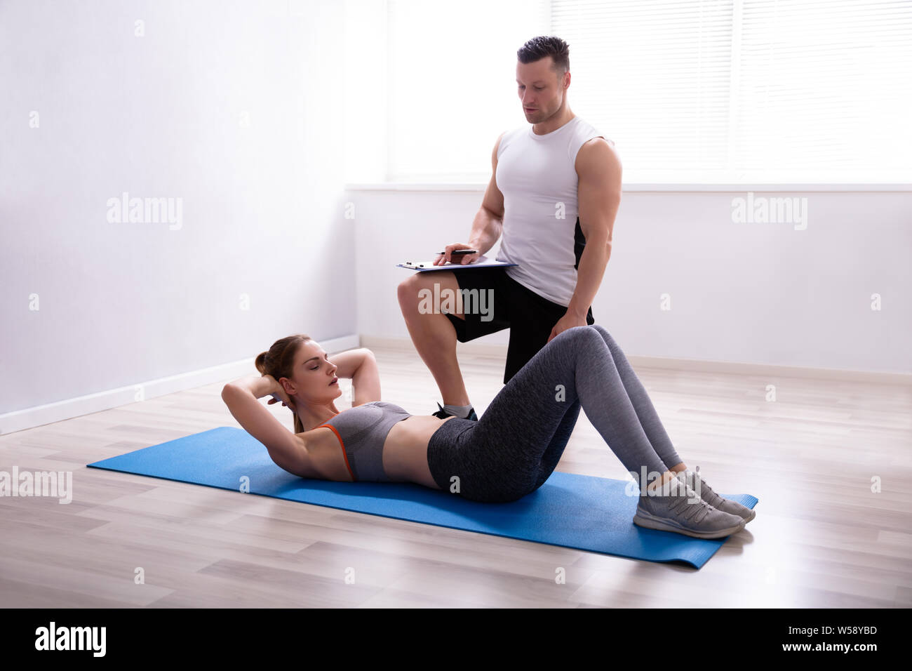 Giovane donna facendo sit ups su materassino yoga accanto al Trainer Holding negli appunti Foto Stock