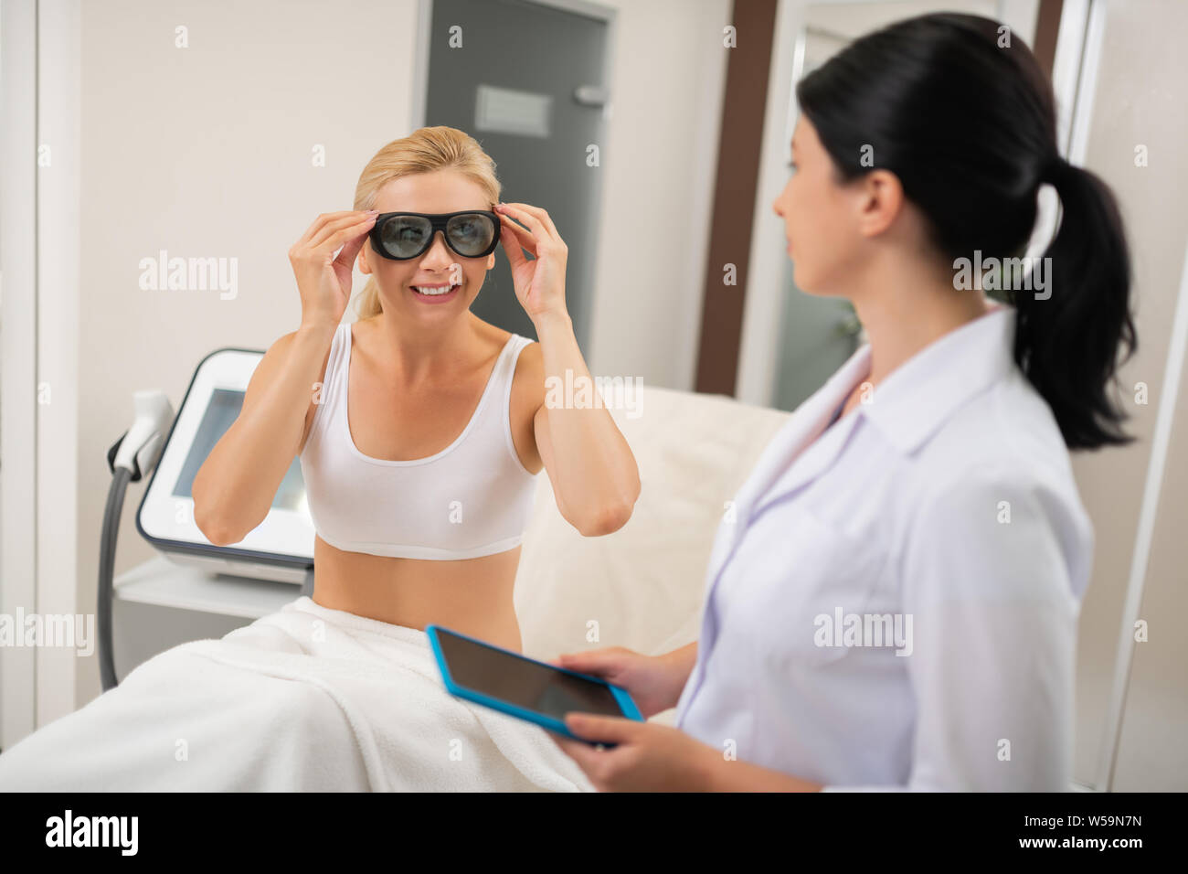 Come rendersi sicuri. Donna sorridente seduto di fronte a lei cosmetologo mettendo su occhiali di protezione prima di procedure. Foto Stock