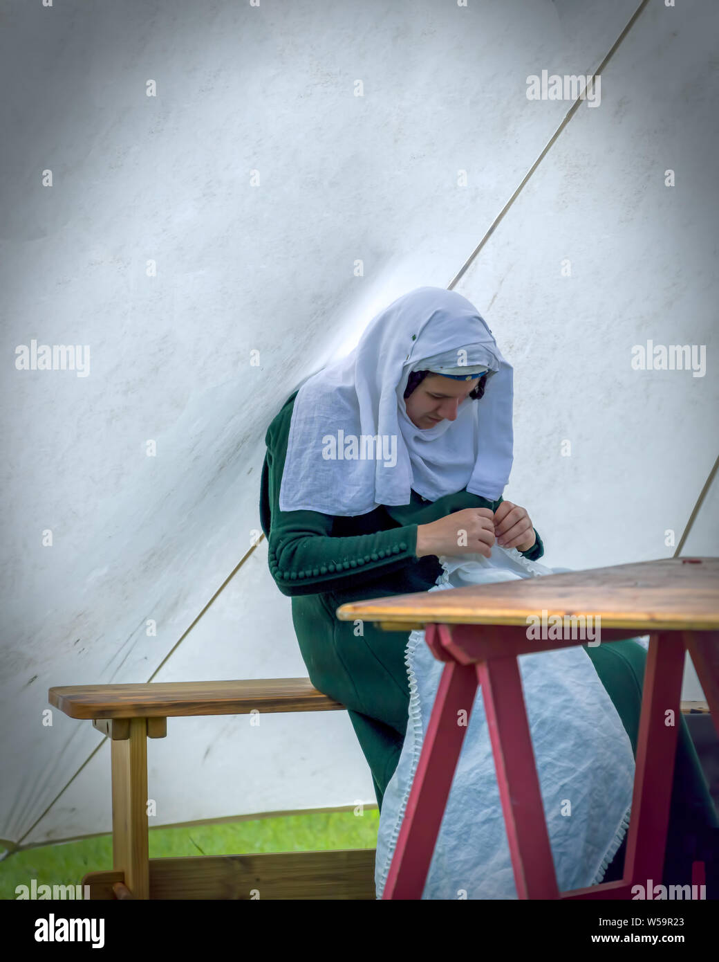 Donna in autentico costume medievale seduto su una panca e tavolo, cucitura in una tenda. Il encampment a Cardiff giostra 2019. Foto Stock