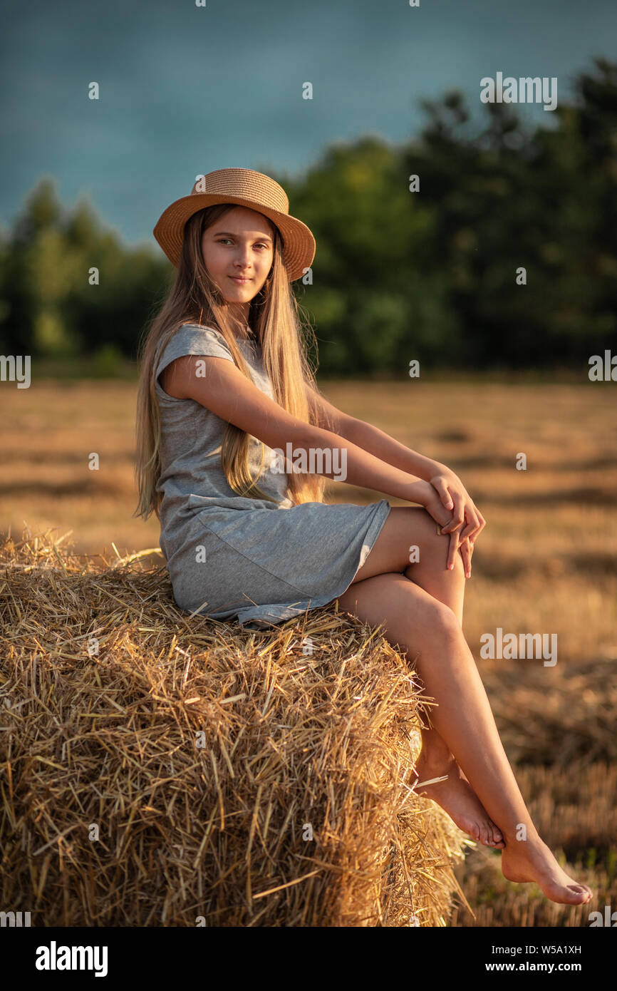 Bella ragazza adolescente con lunghi capelli biondi in un cappello di paglia seduto su di una balla di paglia. Tramonto d'estate. Foto Stock
