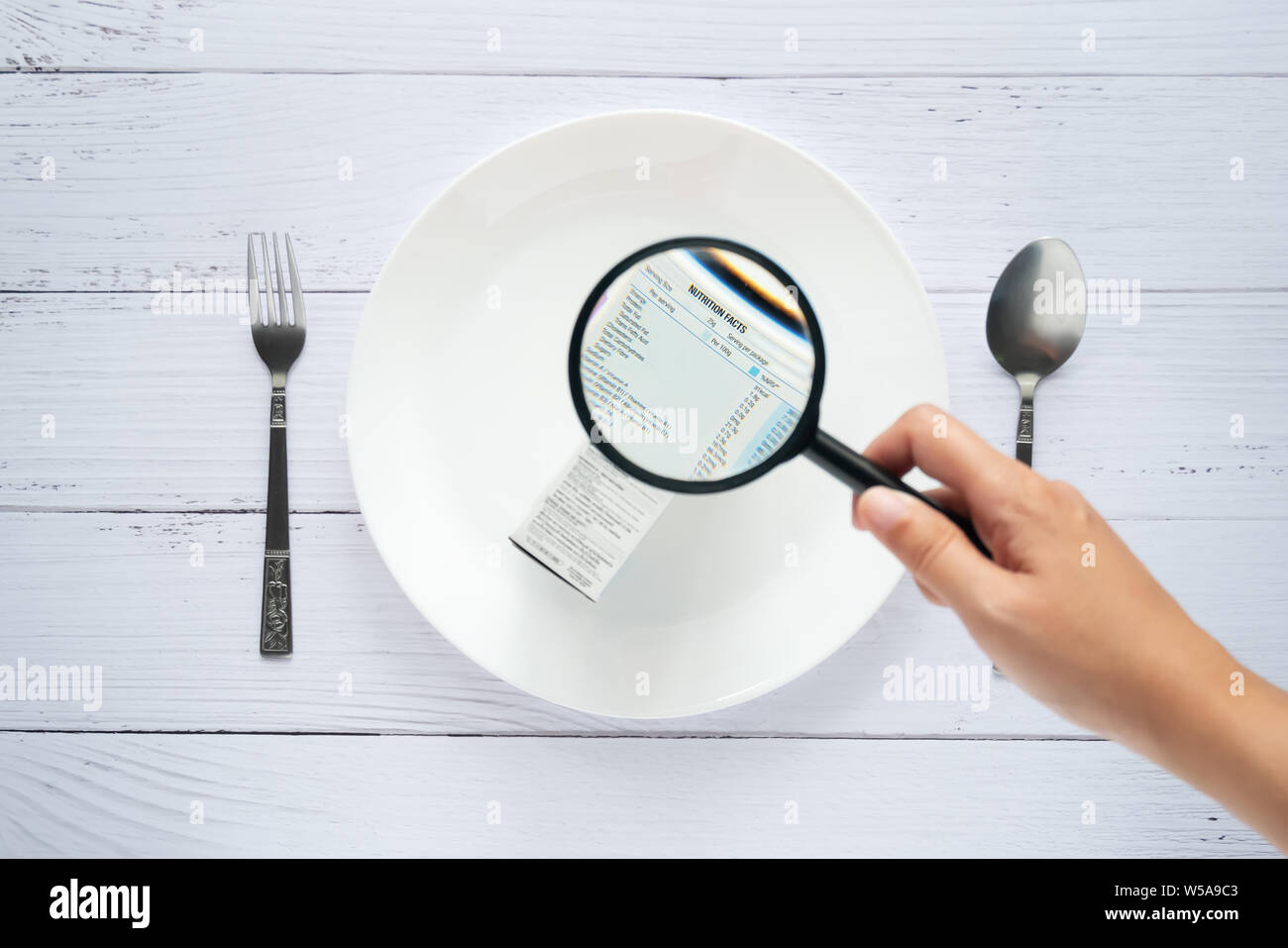 Uso a mano la lente di ingrandimento per ingrandire per vedere i dettagli dei fatti di nutrizione etichetta sul lato del prodotto di consumo casella sul piatto bianco Foto Stock