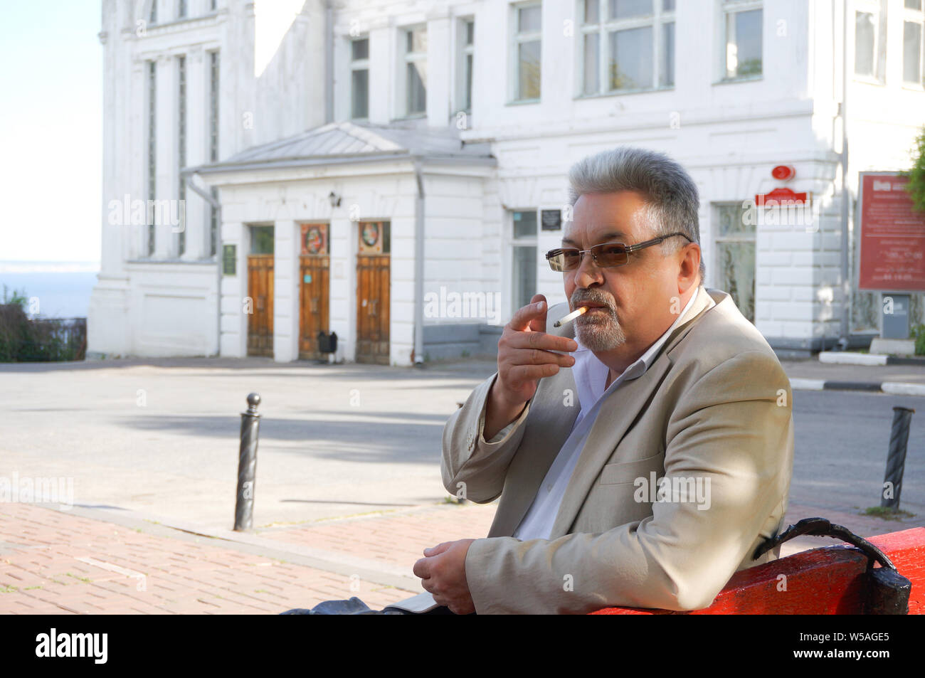 Ritratto di eldery uomo bello con i capelli grigi e bicchieri di fumare mentre è seduto su un banco di lavoro in posizione di parcheggio Foto Stock