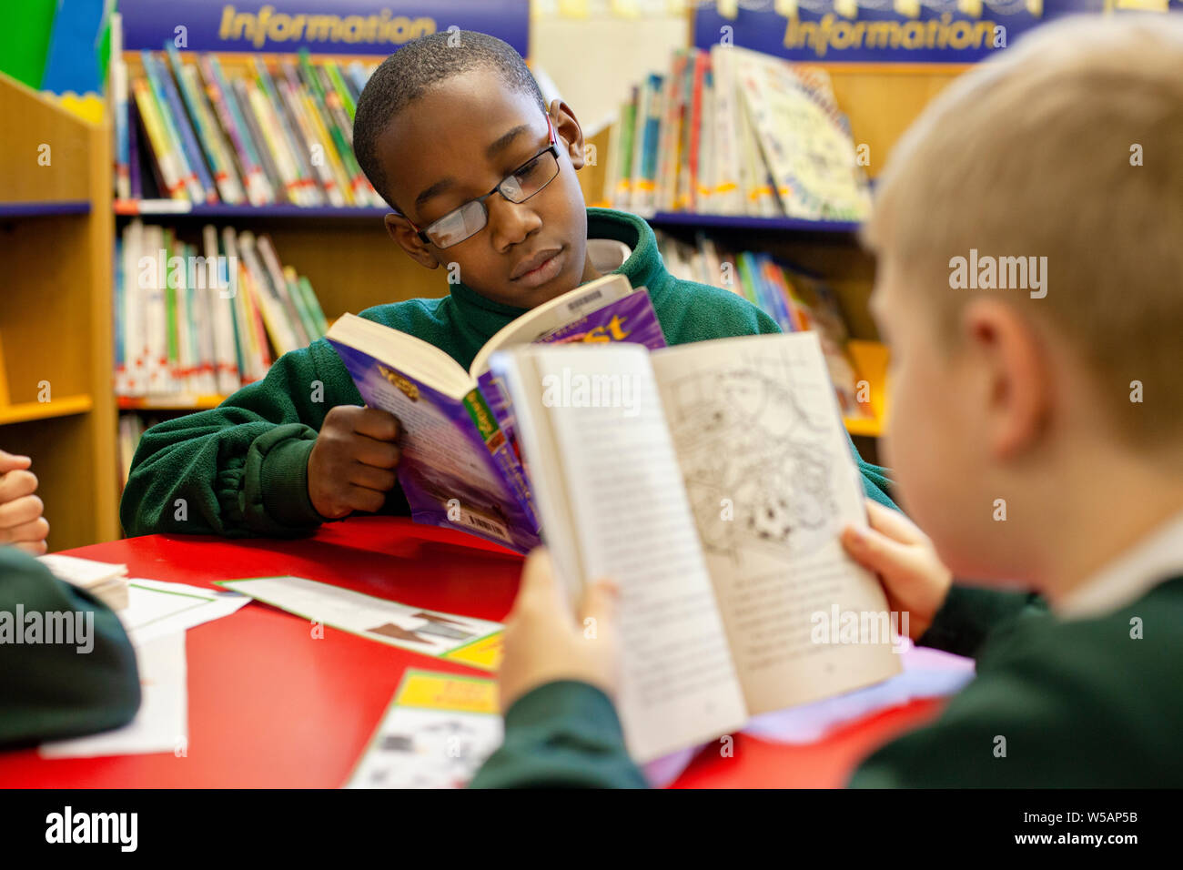 Un ragazzo afro-caraibica e un ragazzo caucasico lettura in biblioteca presso una scuola primaria nel Regno Unito. Foto Stock