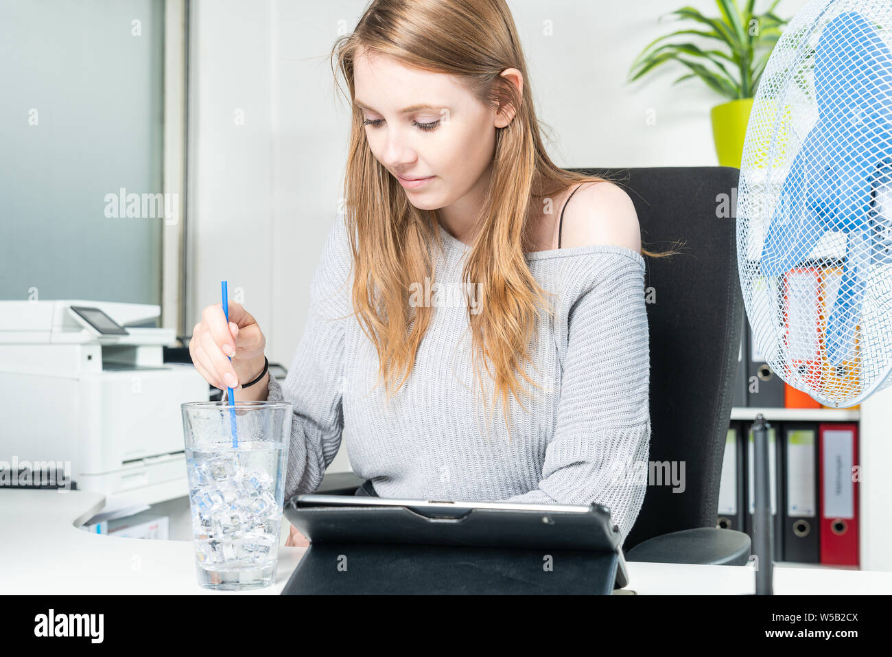 Una giovane donna in ufficio non si raffredda con un bicchiere di acqua pieno di cubetti di ghiaccio Foto Stock