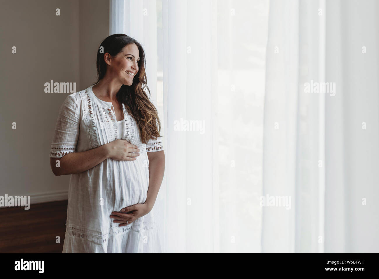 Gravidanza madre sorridente in bianco guardando fuori della finestra di studio Foto Stock