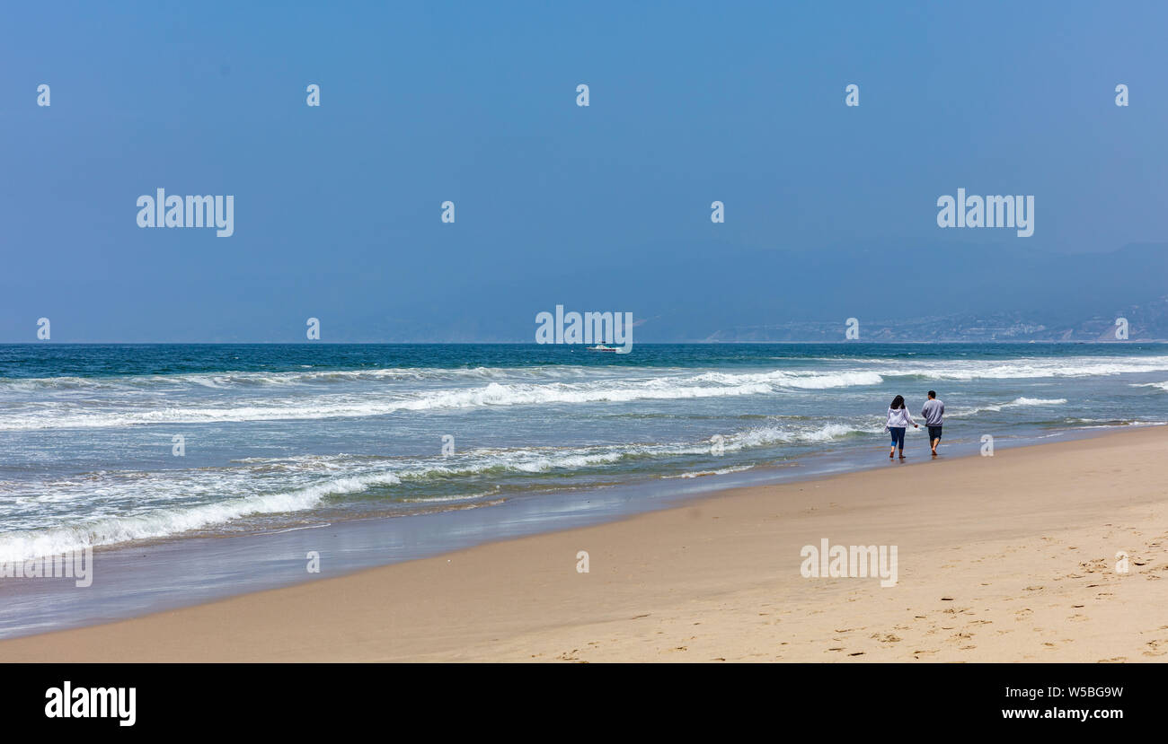 Coppia giovane camminando sulla spiaggia sabbiosa. Blu oceano onde, una barca di salvataggio in mare in una soleggiata giornata di primavera Foto Stock