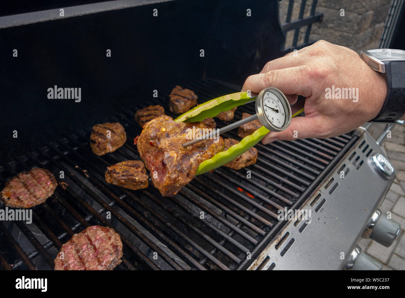 Hamburger e costolette di agnello su un barbecue esterno con grill cucinare mano e alette cheacking le carni di temperatura Foto Stock