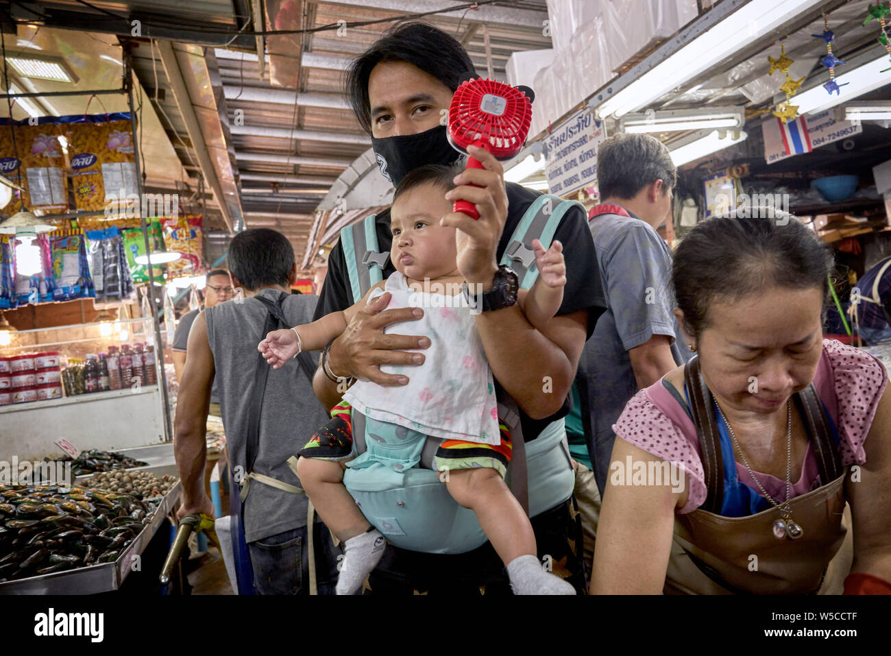 Le persone che fanno caldo si mantengono fresche. Padre che trasporta un bambino e usa un ventilatore portatile per mantenersi fresco nel caldo Tailandia di un mercato interno. Foto Stock
