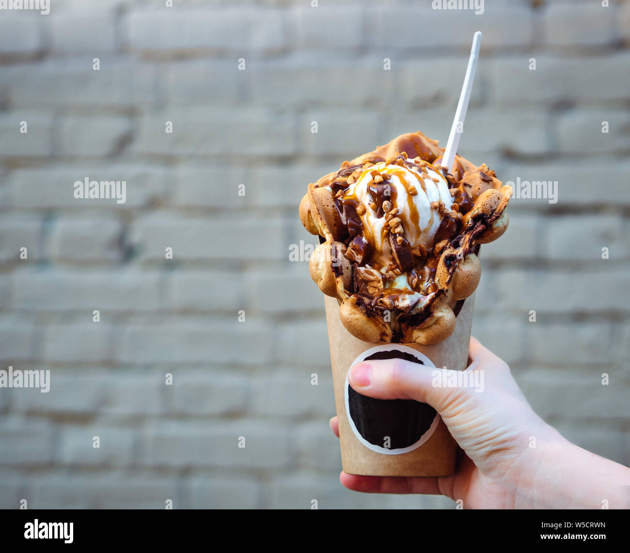 Hong Kong cialde in mano femmina sul muro di mattoni in background. Bolla di deliziosi waffle con gelato e sciroppo di caramello nella donna la mano. Cibo alla moda. Copia spazio per il testo Foto Stock