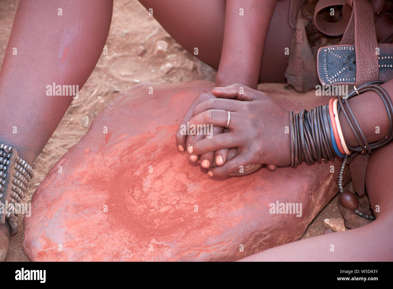 Donna Himba schiaccia l'argilla rossa, questo viene poi utilizzato per rivestire i loro capelli. Fotografato in un villaggio Himba, Epupa Falls, Kaokoland, Namibia, Africa Foto Stock
