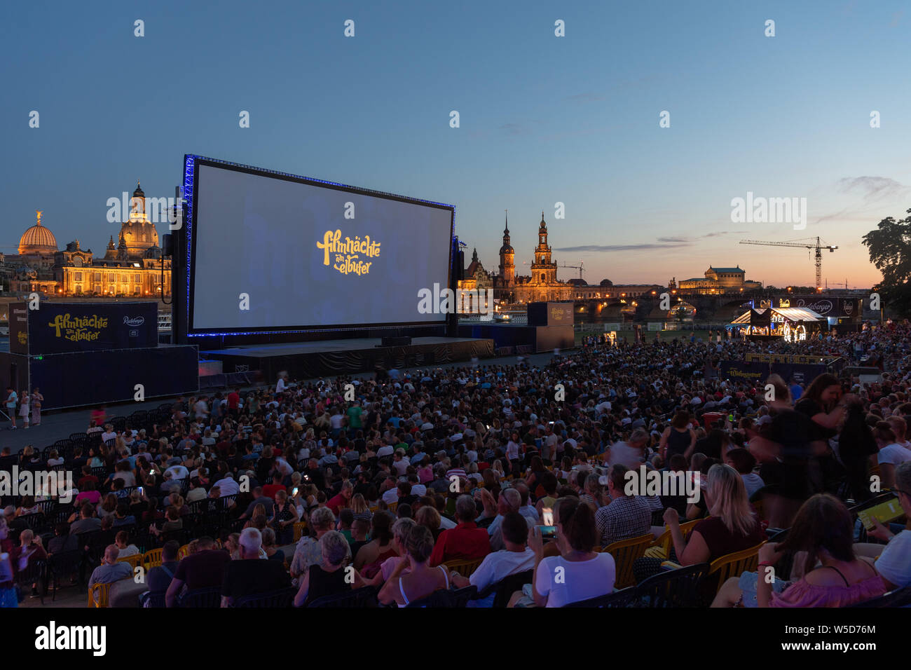 24 luglio 2019, Sassonia, Dresda: numerosi ospiti del 'Filmnächte am Elbufer' sedersi di fronte allo scenario del centro storico di Dresda con la cupola della Kunstakedemie (l-r), la Frauenkirche, la Hausmannsturm, la Hofkirche e il Semperoper e guardare uno schermo cinematografico. Foto: Robert Michael/dpa-Zentralbild/dpa Foto Stock