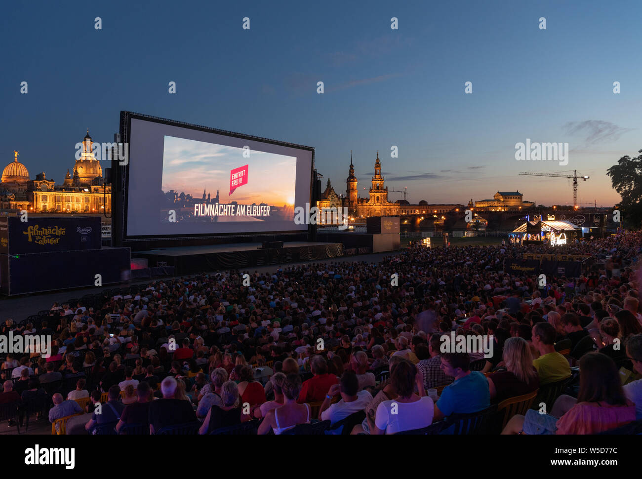 24 luglio 2019, Sassonia, Dresda: numerosi ospiti del 'Filmnächte am Elbufer' sedersi di fronte allo scenario del centro storico di Dresda con la cupola della Kunstakedemie (l-r), la Frauenkirche, la Hausmannsturm, la Hofkirche e il Semperoper e guardare uno schermo cinematografico. Foto: Robert Michael/dpa-Zentralbild/dpa Foto Stock