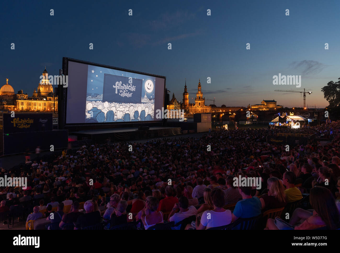 24 luglio 2019, Sassonia, Dresda: numerosi ospiti del 'Filmnächte am Elbufer' sedersi di fronte allo scenario del centro storico di Dresda con la cupola della Kunstakedemie (l-r), la Frauenkirche, la Hausmannsturm, la Hofkirche e il Semperoper e guardare uno schermo cinematografico. Foto: Robert Michael/dpa-Zentralbild/dpa Foto Stock