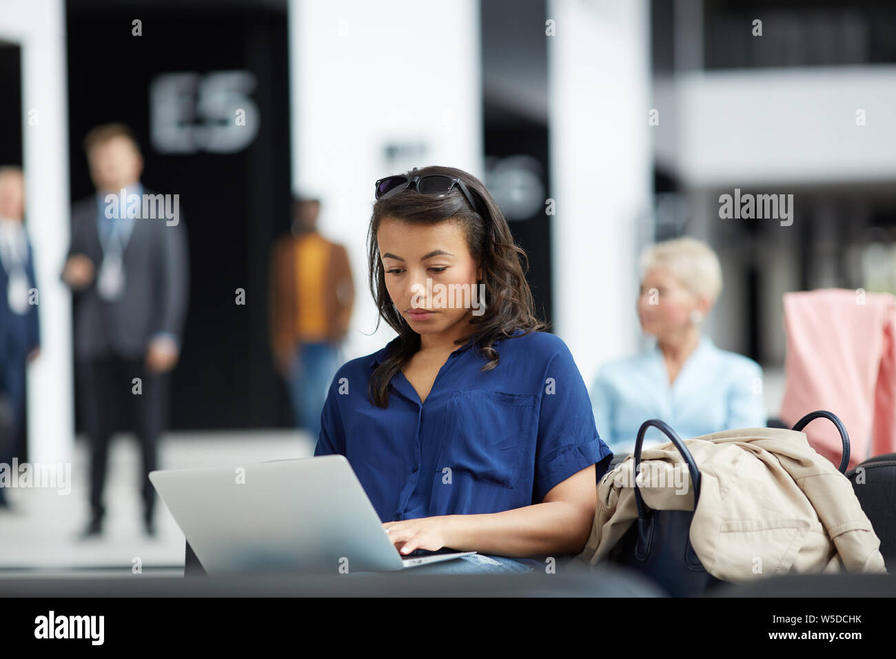 Grave occupato giovane nero donna con occhiali da sole sulla testa seduto sul divano in aeroporto e digitando su laptop Foto Stock