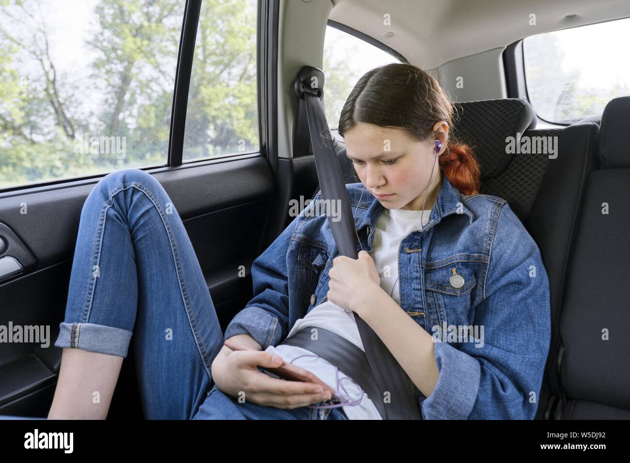 Teen ragazza seduta in auto nel retro del sedile passeggero con lo smartphone e le cuffie. Foto Stock