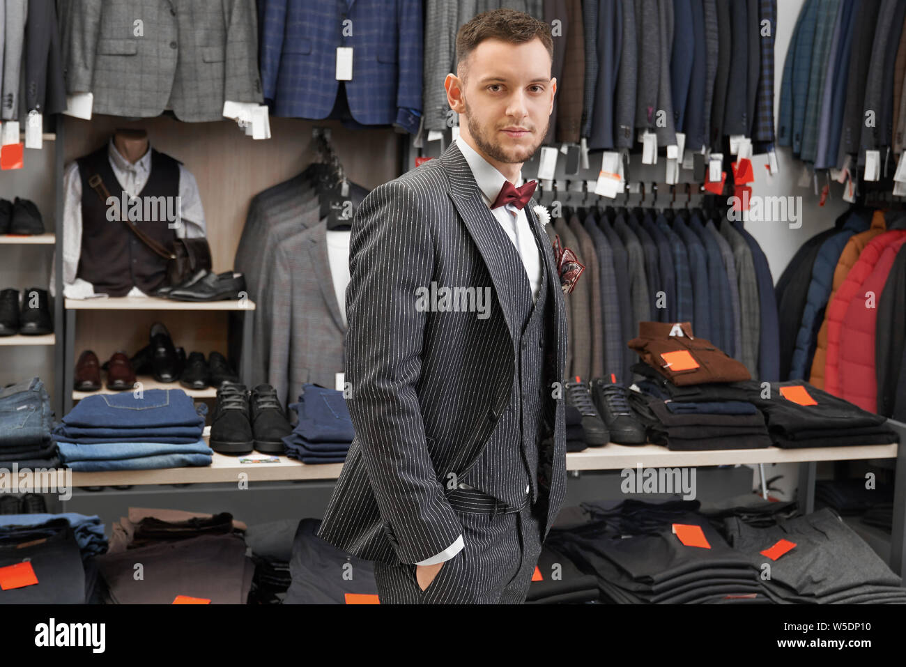 Giovani, fiducioso uomo con barba elegante in posa elegante abito, gilet, una camicia bianca e rosso il filtro bow tie. Bello, imprenditore di successo in piedi, tenendo le mani nelle tasche, guardando la fotocamera. Foto Stock