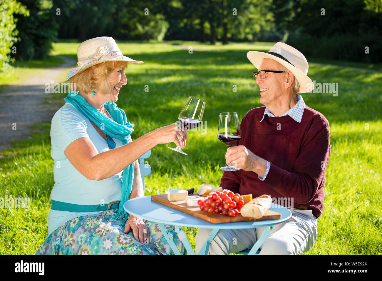 Coppia senior in amore fare picnic nel parco, seduti a un tavolo che ognuno guarda negli occhi dell'altro, bere vino e mangiare il formaggio con baguette e i mosti di uve Foto Stock