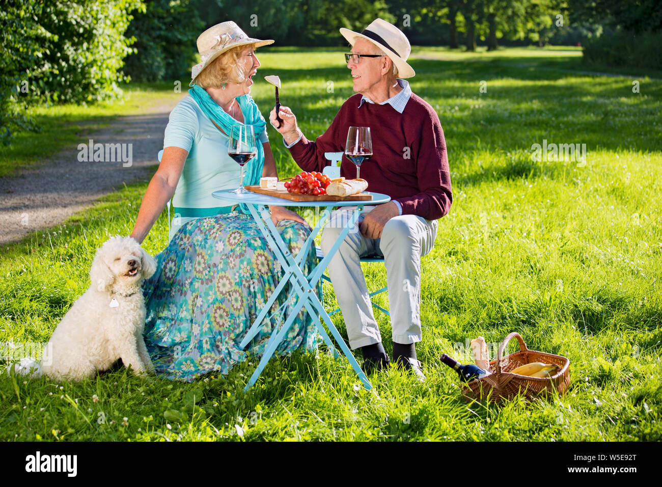 Felice coppia senior con un cane bianco picnic nel parco. Bionda bella donna anziana è aprire la bocca e il suo fidanzato è la sua alimentazione con Foto Stock