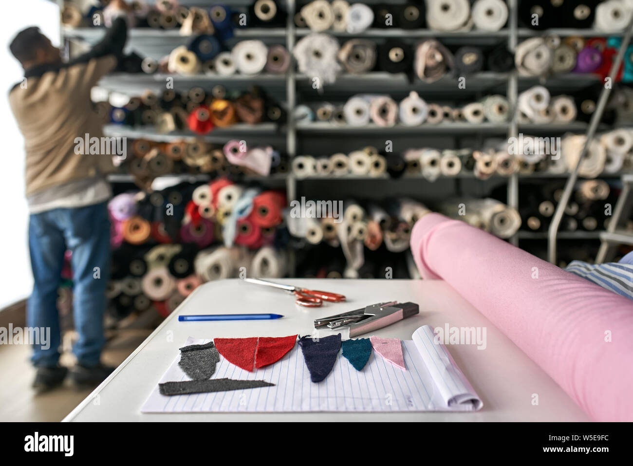 Uomo sceglie un materiale da ripiani con multicolore di rotoli di tessuto all'interno di un magazzino. Davanti a lui non c è tavolo bianco con pezzi di tessili e ac Foto Stock
