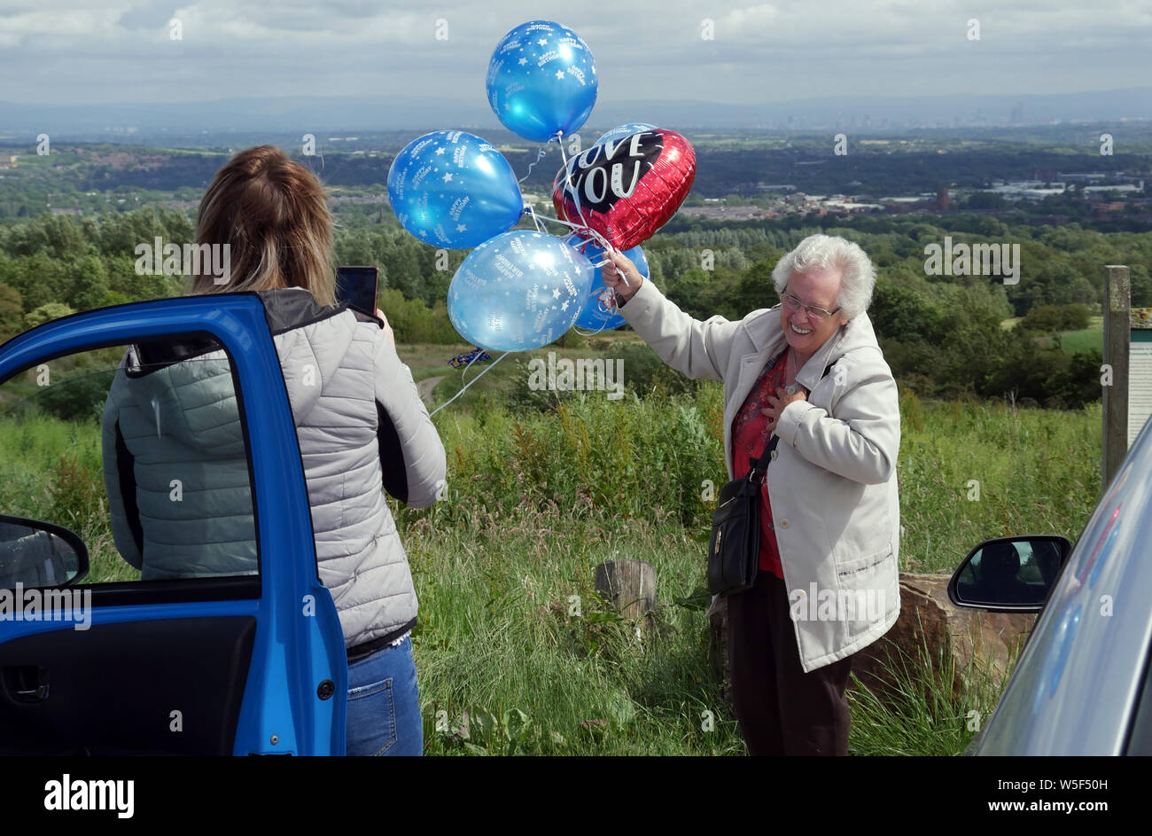 Signora anziana il rilascio di palloncini, un palloncino è a forma di cuore dicendo "ti amo" come una giovane donna registra l'evento con il suo smart phone su un parco auto Foto Stock