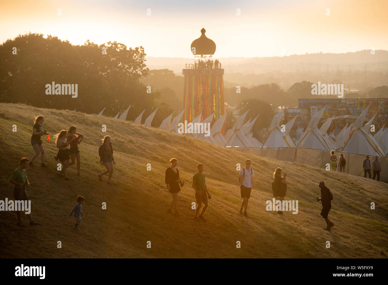 I frequentatori del festival al di sopra del villaggio di tende Tepee e Torre a nastro come il sole tramonta al Glastonbury festival 2019 in Pilton, Somerset Foto Stock