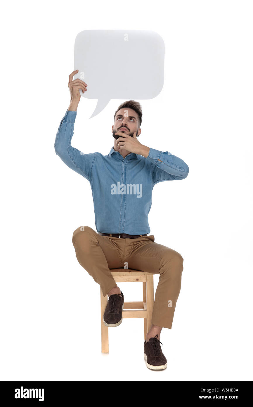 Seduto curioso uomo in blue jeans shirt pensando mentre tiene un vuoto parlare di cloud in aria su sfondo bianco Foto Stock