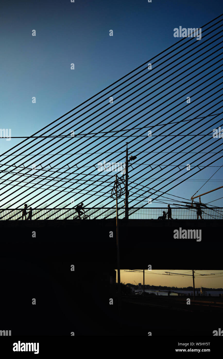 Diverse sagome di persone andare e un giro in bici su un cavo-alloggiato ponte con fili in Riga al tramonto Foto Stock