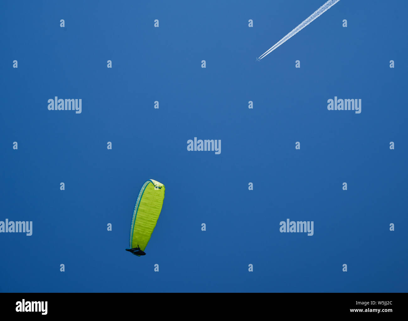 Un parapendio con un giallo-verde parashute e un aereo lasciando un sentiero bianco sul cielo blu. Foto Stock