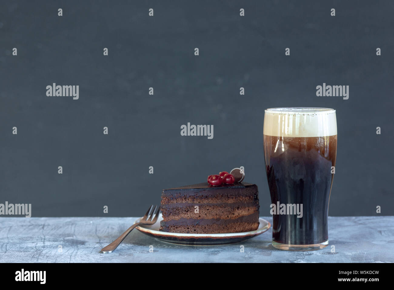 Bicchiere di birra scura sul tavolo di pietra e uno sfondo grigio. Bevanda fredda e una fetta di torta o dolci sono preparati per una grande festa. Concetto di bevande, divertente, cibo, celebrando, sale riunioni, Oktoberfest. Foto Stock