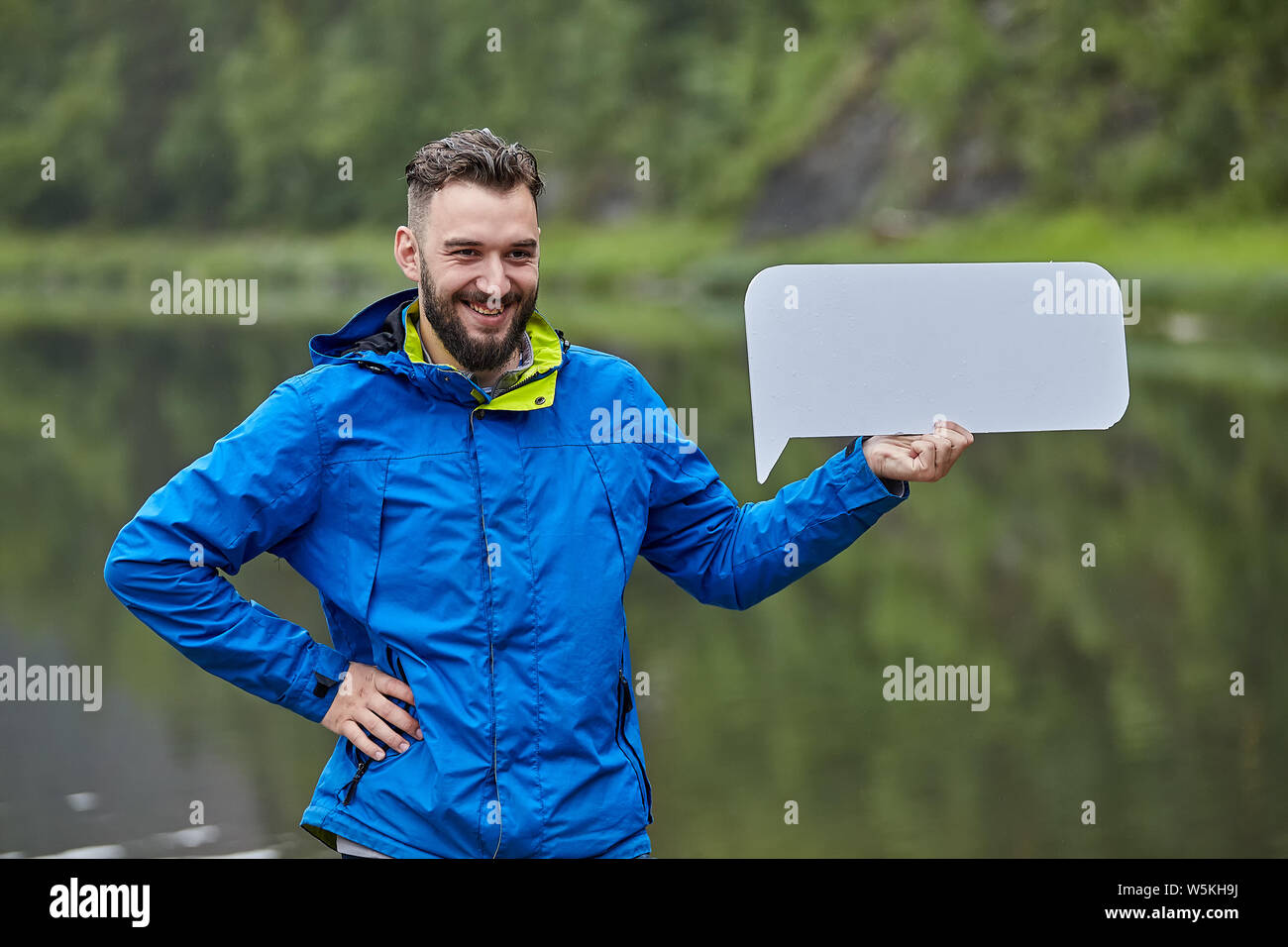Giovani attraenti barbuto uomo caucasico con foglio bianco è in piedi nella natura selvaggia vicino al fiume in giacca blu da soli e sorridente. Foto Stock