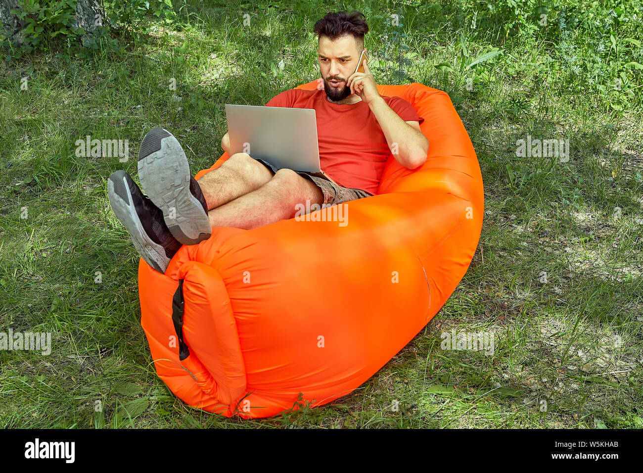 Giovane imprenditore barbuto è sdraiato sul divano aria sull'erba e lavorare usando il suo computer portatile e parlare al telefono durante l'eco-turismo. Foto Stock
