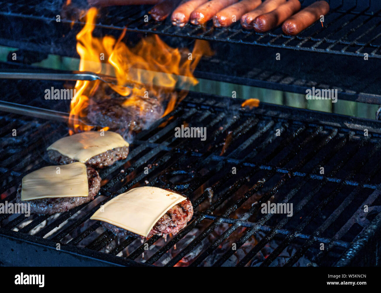Pinze sono di prelevare un hamburger come fiamme dal carbone che lo circondano e cheeseburger e hot dogs sono tutti cottura alla griglia. Foto Stock