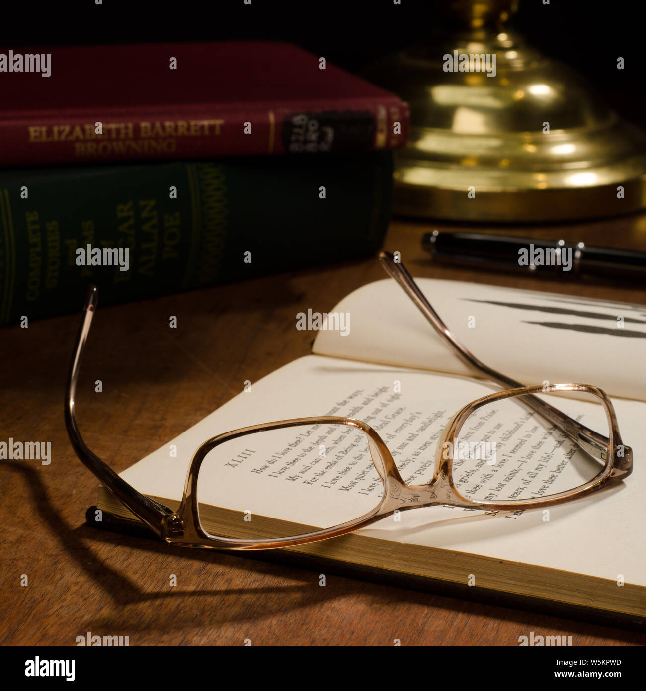 Un paio di occhiali di cui capovolto su un libro aperto, con altri libri e una penna a inchiostro su di una scrivania con lampada in ottone. Foto Stock