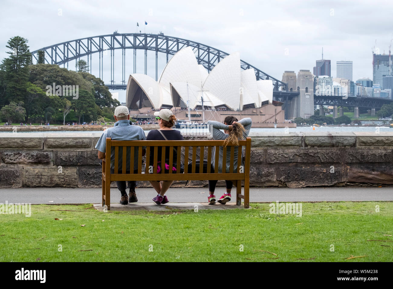 Tre persone a sedersi su una panchina a guardare fuori per la Sydney Opera House di Sydney e il Sydney Harbour Bridge dalla fattoria Cove, Sydney Foto Stock