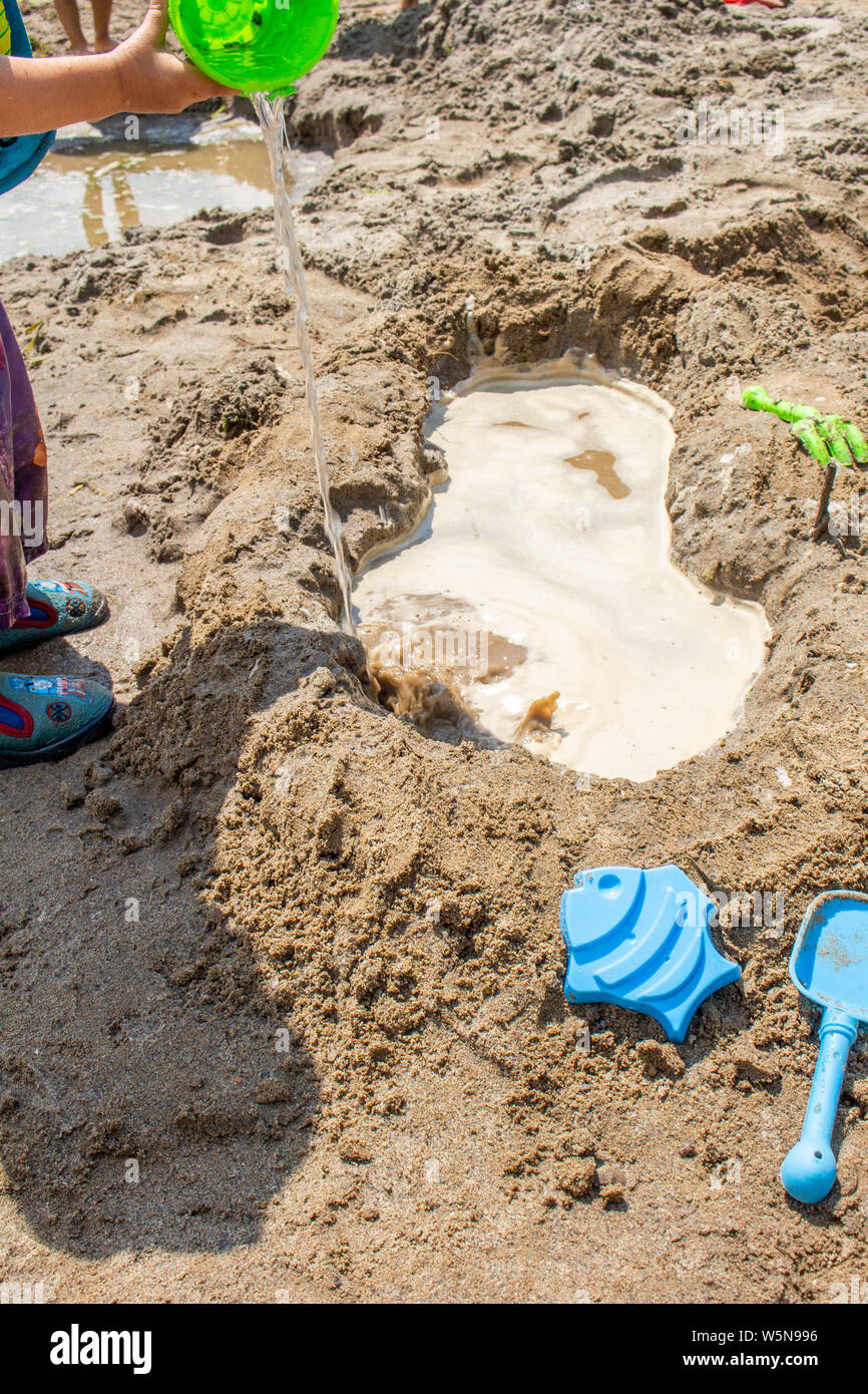 Un giovane bambino sorge in corrispondenza del bordo della cornice, versando un secchio di acqua in un foro scavato in una spiaggia sabbiosa, con sabbia giocattoli disseminate attorno ad esso. Foto Stock