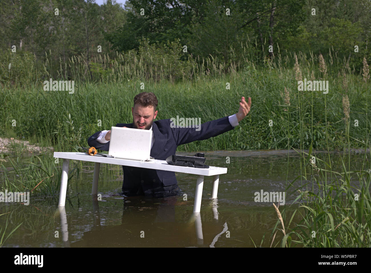 Giovani barbuto uomo millenario, guy stanco del lavoro d'ufficio. Arrabbiato e distratto sul posto di lavoro. A tavola in piedi la palude, in acqua Foto Stock