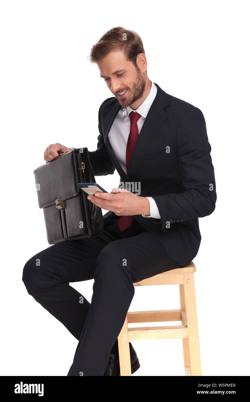 Seduto imprenditore tenendo la valigia è in attesa per l'intervista e scrivere messaggi su telefono cellulare su sfondo bianco Foto Stock